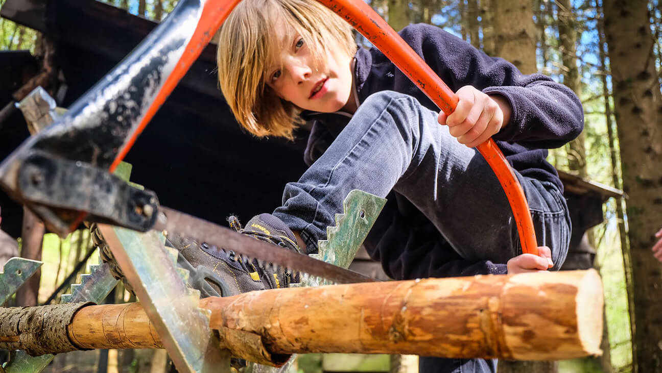 Jongen zaagt houtblok tijdens kamp van Outdoorschool