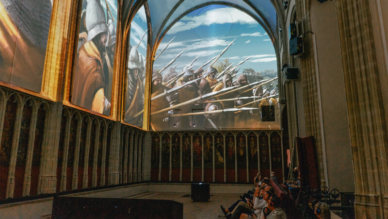 Mensen kijken naar groot scherm op de tentoonstelling '1302' in de Onze-Lieve-Vrouwekerk in Kortrijk