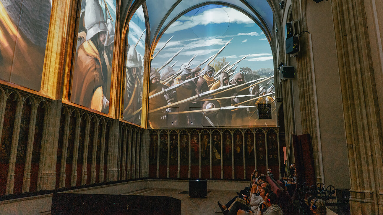 Mensen kijken naar groot scherm op de tentoonstelling 1302 in de Onze-Lieve-Vrouwekerk in Kortrijk