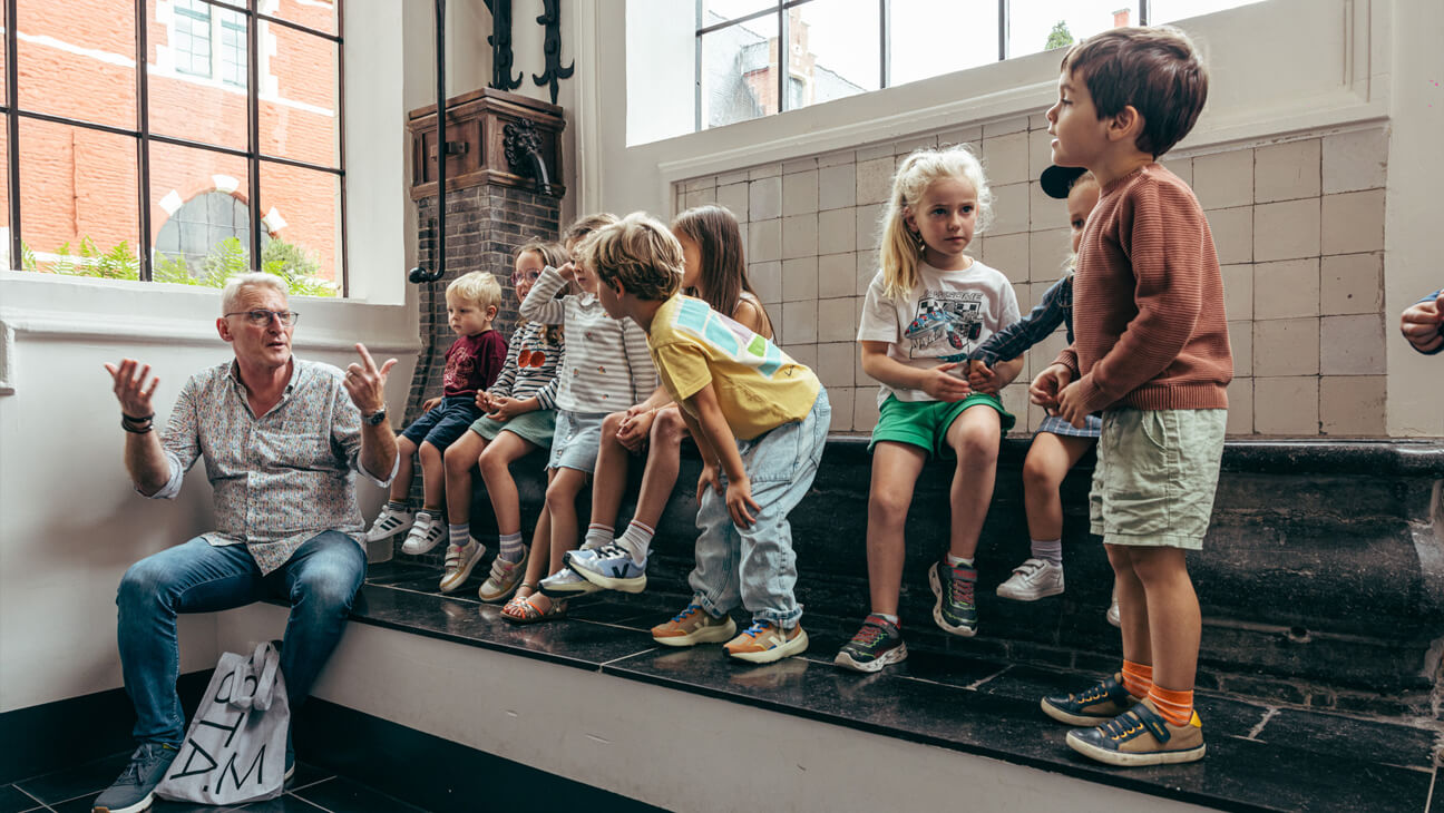 Groep kinderen zitten op een verhoog en luisteren naar een gids