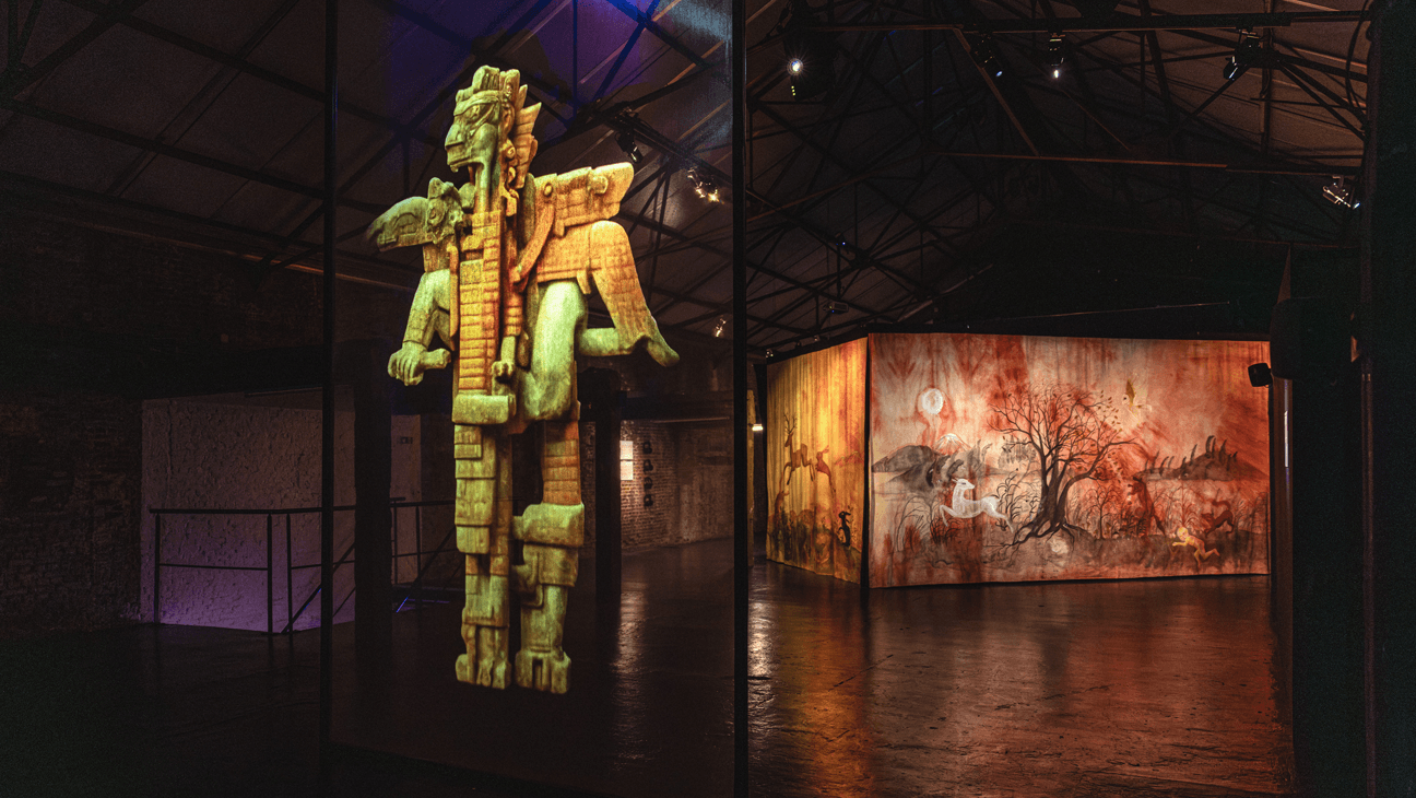 Museumzaal met op een groot scherm een aztekenbeeld, in de achtergrond een groot scherm met hiërogliefen