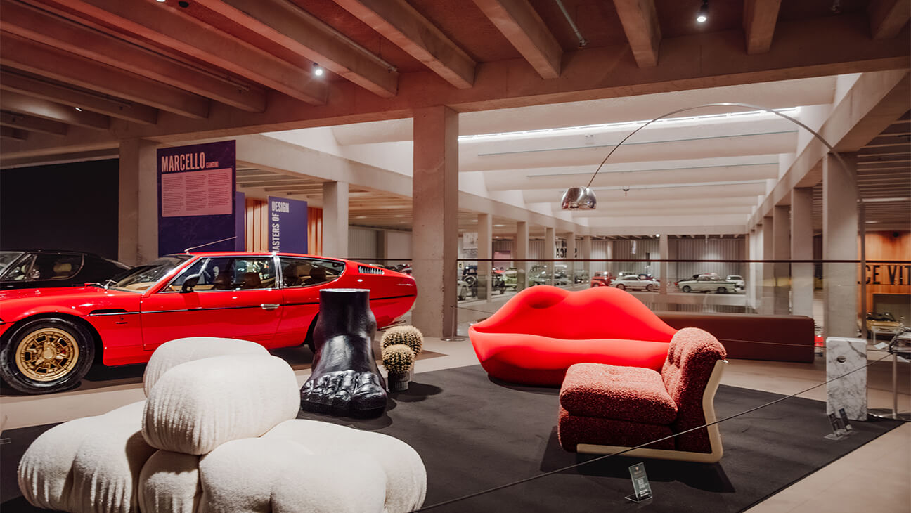 Zaal in een museum met designmeubels met op de achtergrond een rode sportwagen